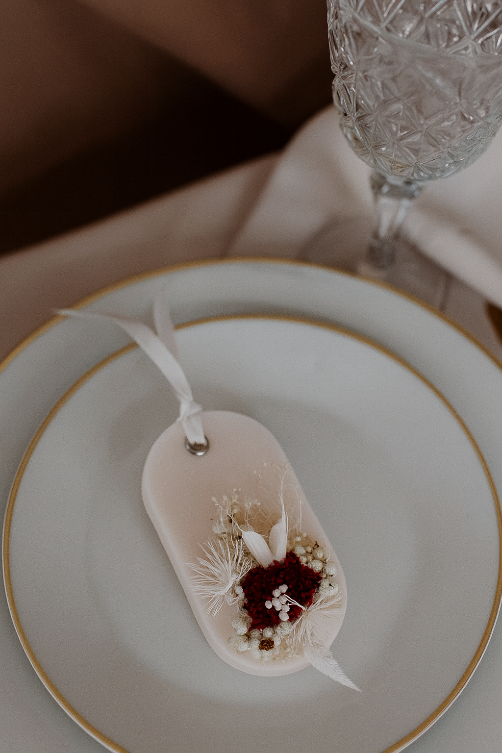Decorative sachet favor with flowers on a white plate with gold rim, next to a crystal glass.