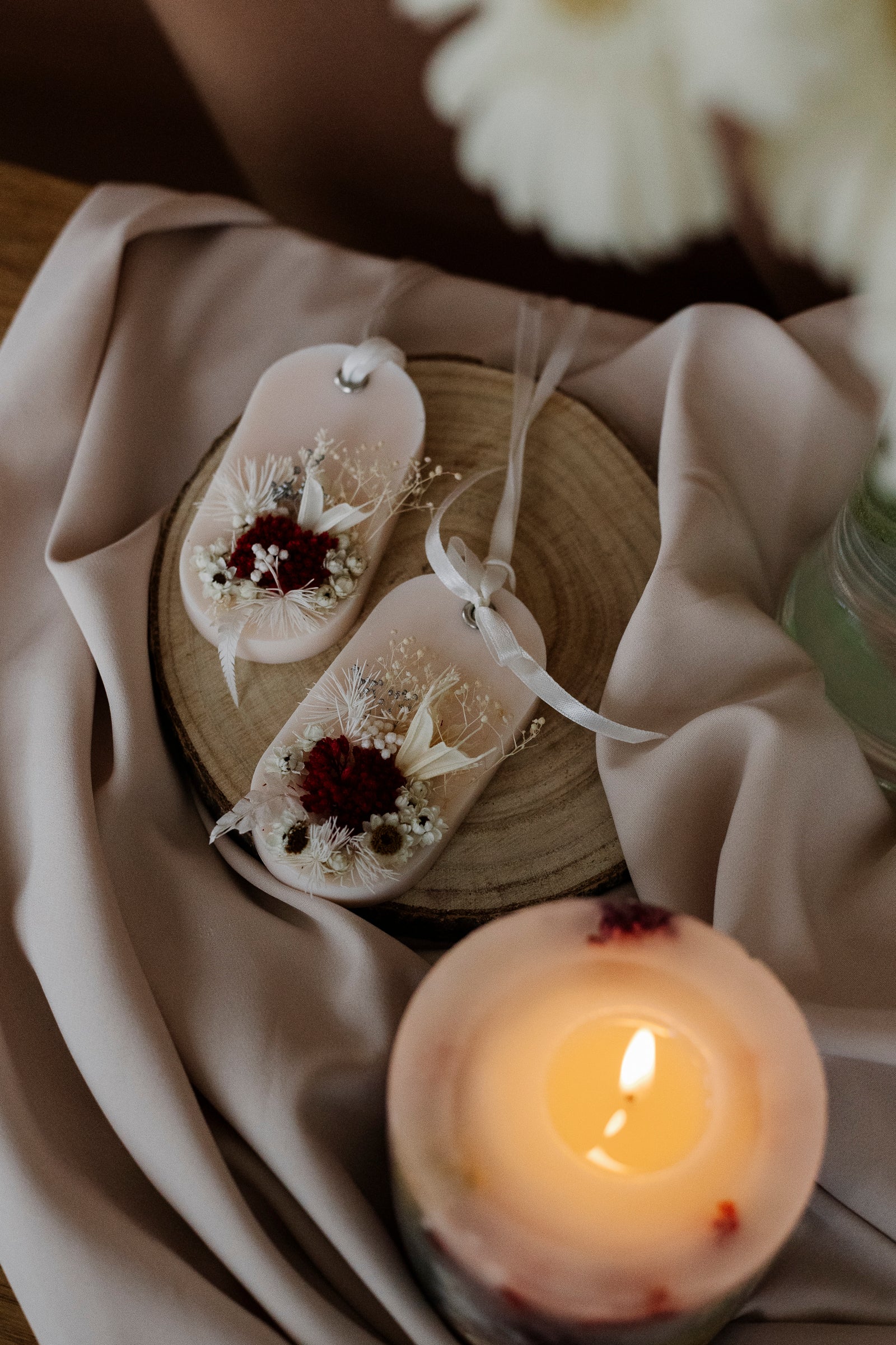 Decorative sachets with flowers on a wooden block next to a lit candle on a soft fabric background