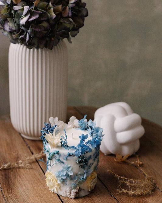 Decorative candle with floral elements on a wooden surface next to a white vase and a white sculpture.