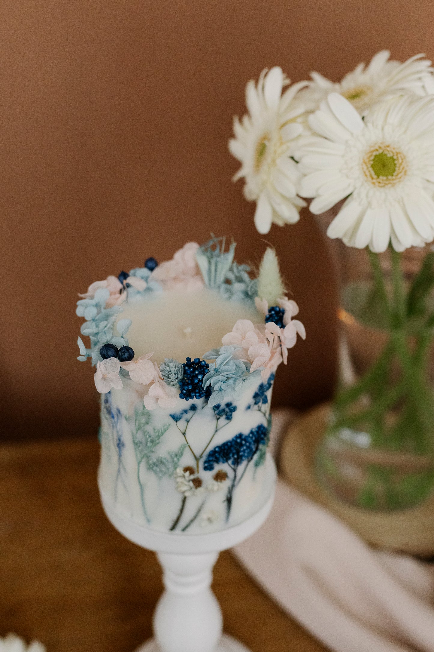 Decorative candle with floral elements on a white stand, with a blurred background of flowers and a brown surface.