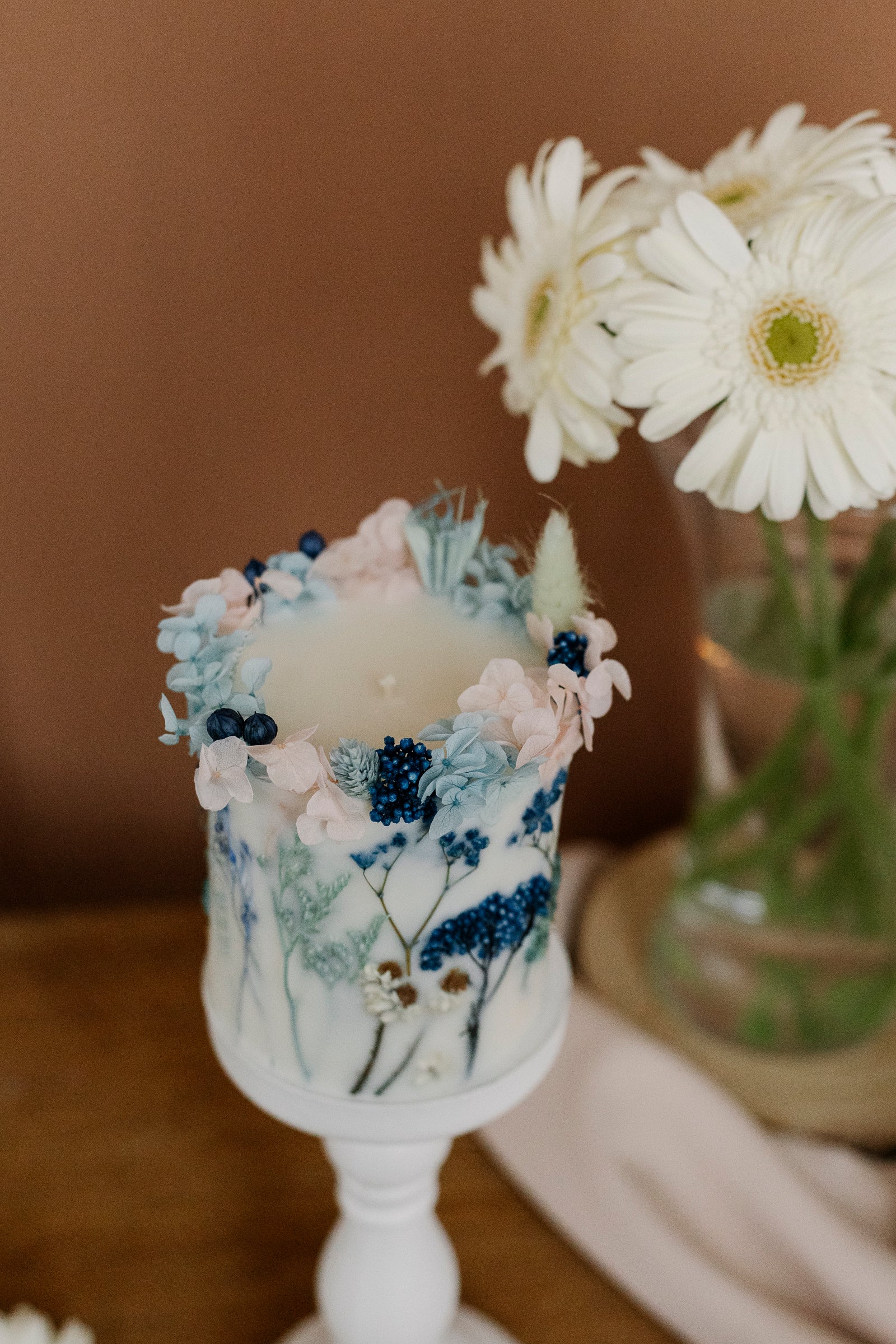 Decorative candle with floral elements on a white stand, with a blurred background of flowers and a brown surface.
