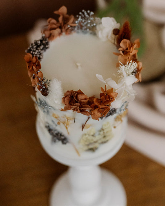 Decorative candle with floral and pinecone arrangement on a white pedestal