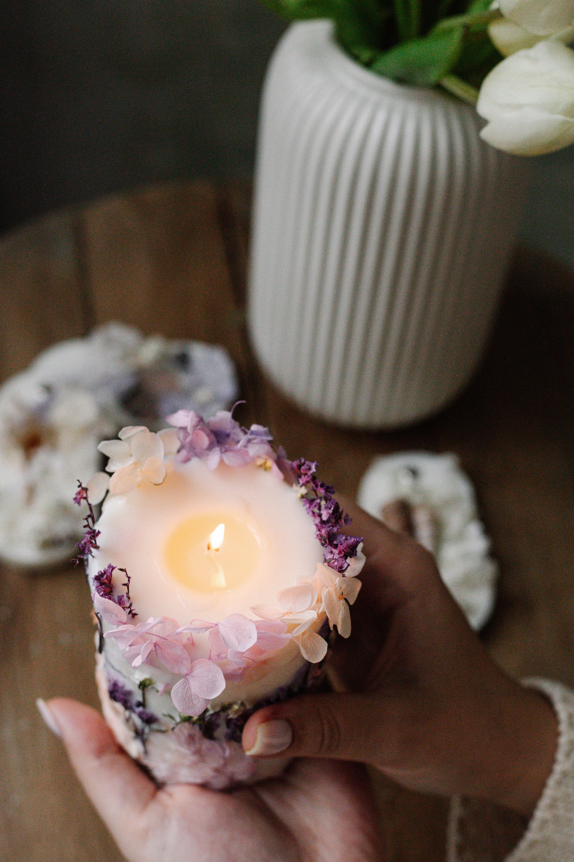 Hand holding a lit candle with flowers, surrounded by a vase of white flowers and decorative stones on a wooden surface.