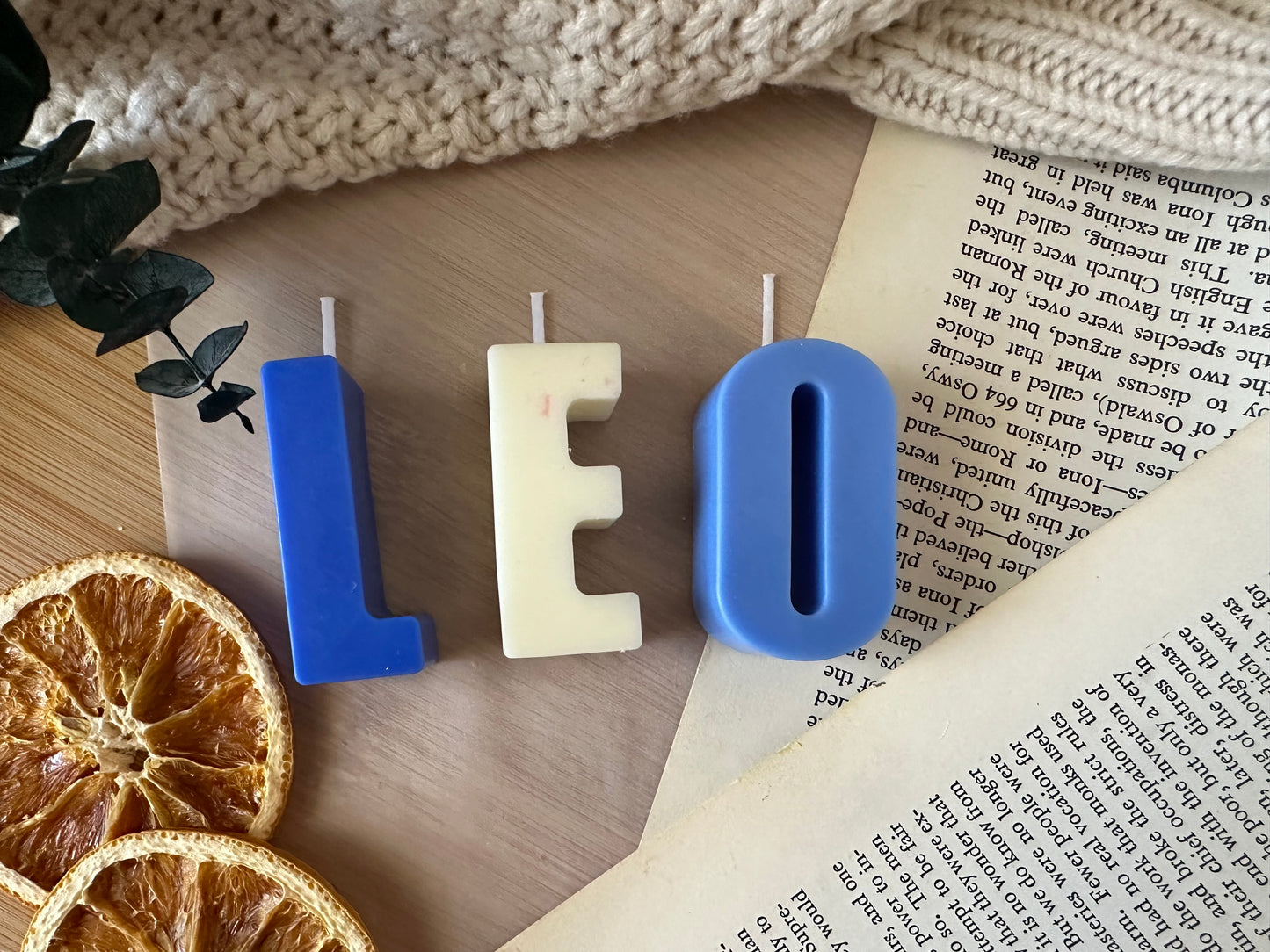 Three letter candles spelling 'LEO' on a wooden surface with dried oranges and a book.