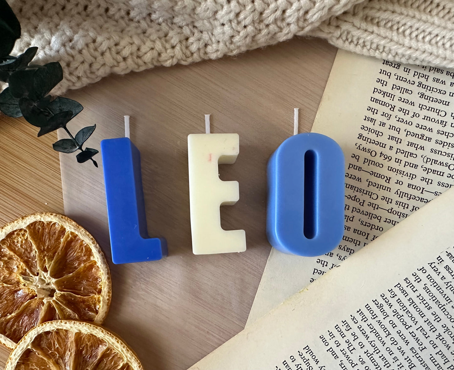 Letter-shaped candles spelling 'LEO' on a wooden surface with dried oranges and a book.