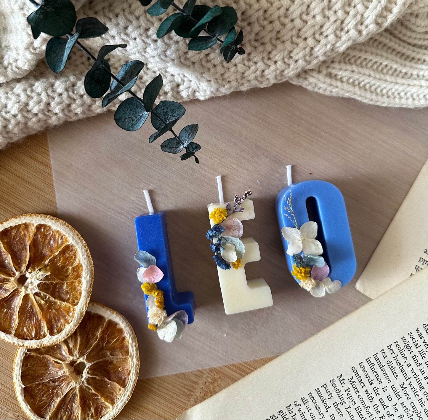 Decorative letter candles with floral elements on a wooden surface with dried oranges and eucalyptus.