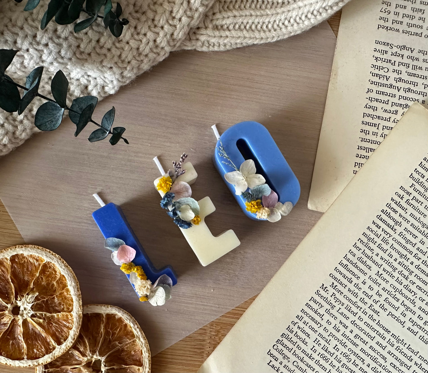 Decorative letter candles 'LEO' with floral elements on a wooden surface next to dried oranges and a book.