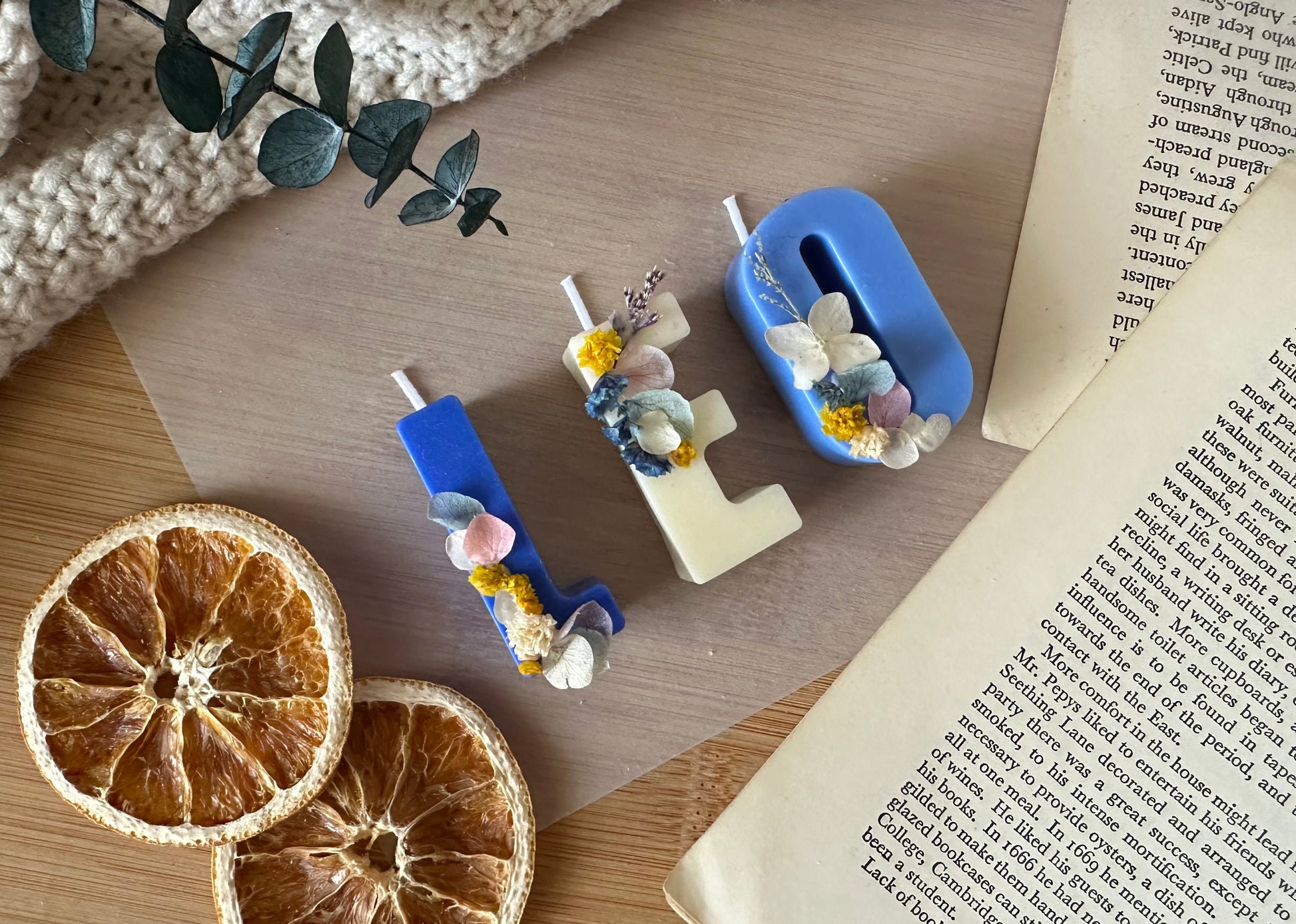 Decorative letter candles 'LEO' with floral embellishments on a wooden surface with dried oranges and a book.