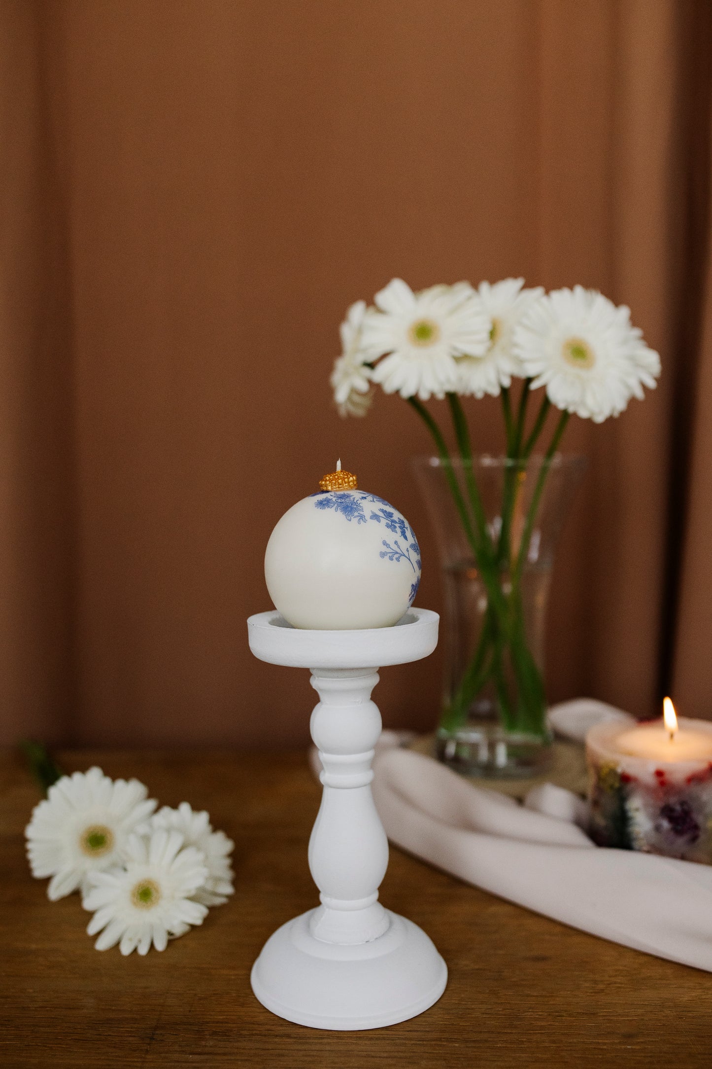 Decorative egg with floral design on a white candlestick, surrounded by flowers and a candle.