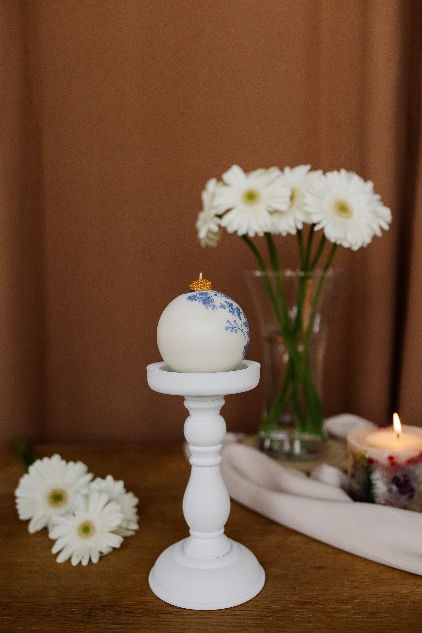 Decorative egg with floral design on a white candlestick, surrounded by flowers and a candle.