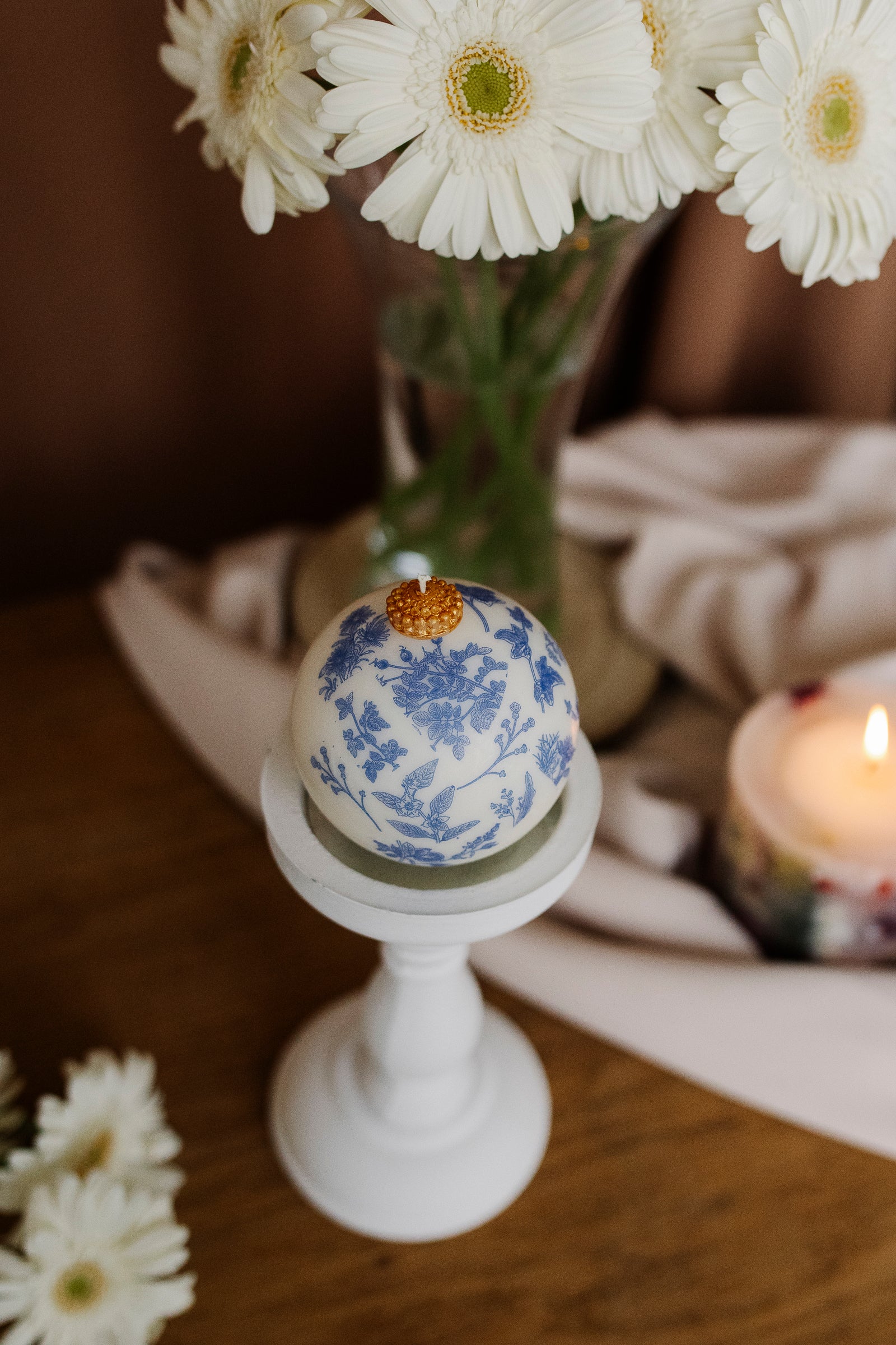 Decorative egg with floral pattern on a white stand, surrounded by white flowers and a candle.