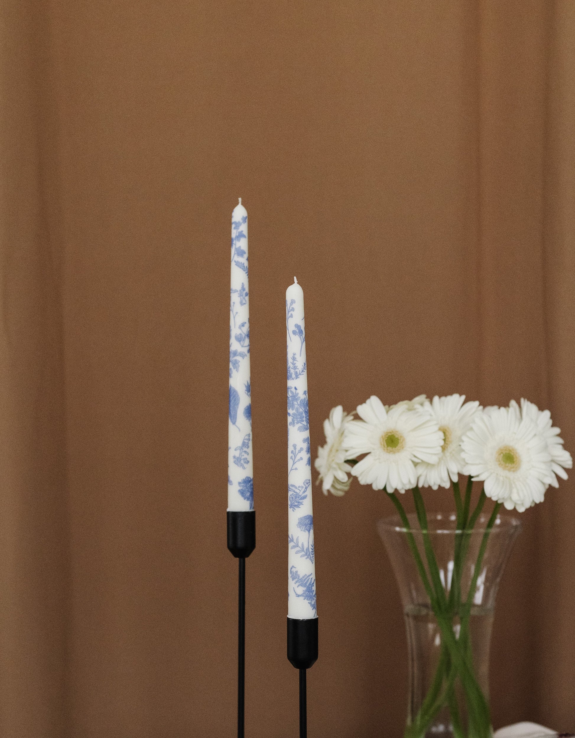 Two floral-patterned candles with black bases on a brown background, accompanied by a vase of white flowers.
