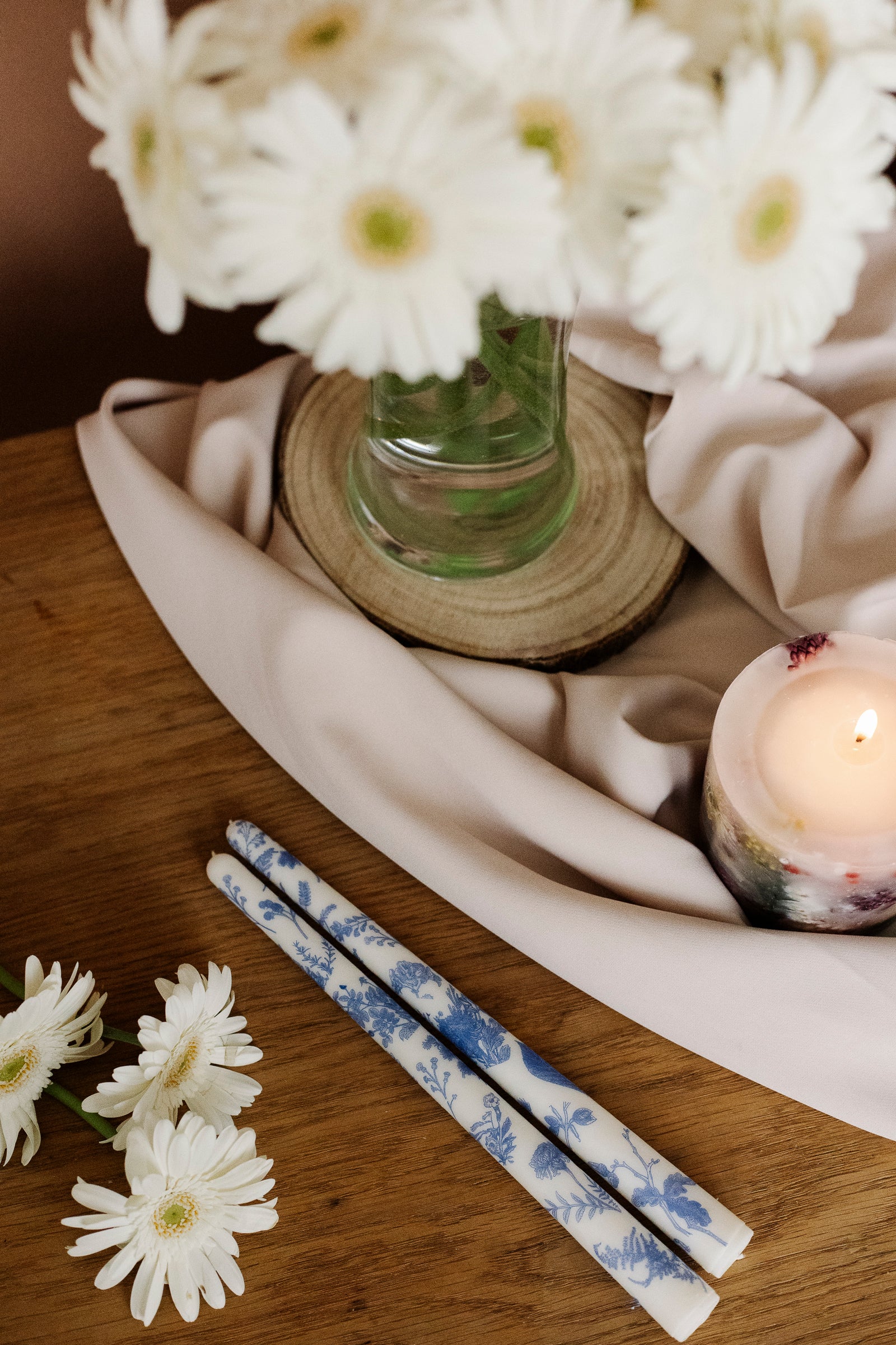 White flowers in a vase on a wooden table with floral-patterned candles and a white figurine.
