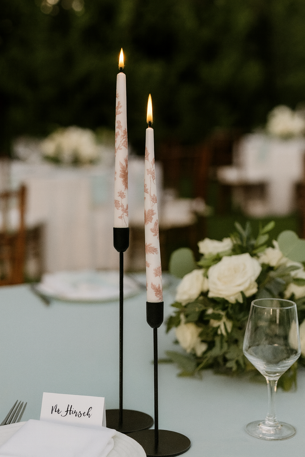 Decorative candles on a table with flowers and a glass in the background