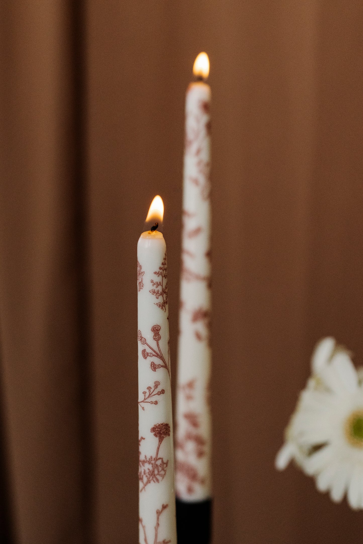 Two white candles with floral patterns and flames against a brown background