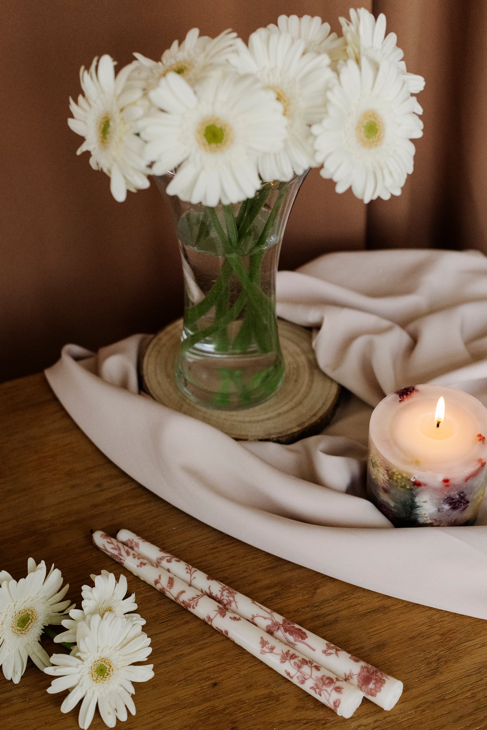 White flowers in a vase on a wooden surface with candles and fabric