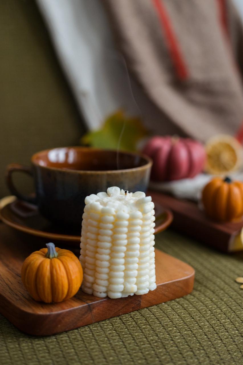 Decorative corn candke on a wooden board with pumpkin candle and a cup in the background