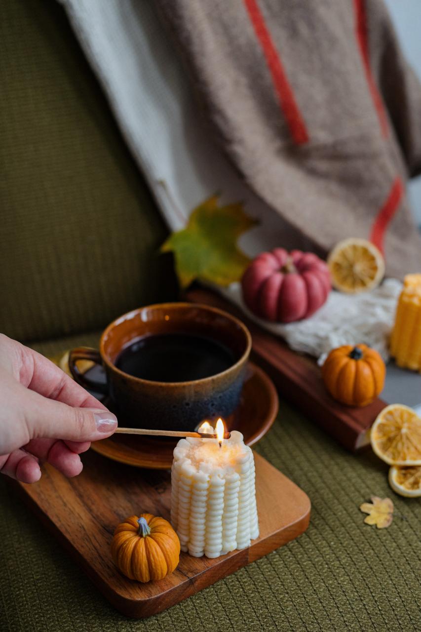 Person lighting a candle with a match, surrounded by pumpkins and a cup of coffee on a wooden tray.
